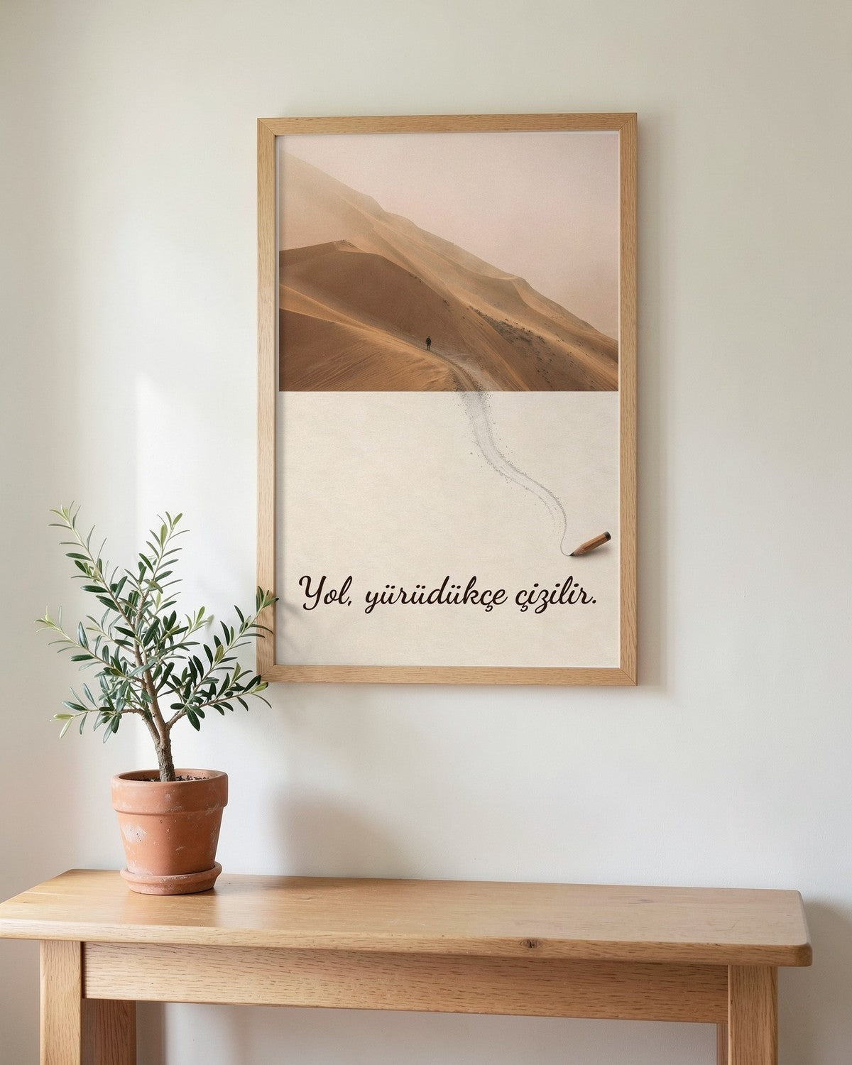 Ahşap çerçeveli çöl ve karakalem poster, açık renk duvarda, saksıda zeytin fidanı olan ahşap konsolun üstünde.