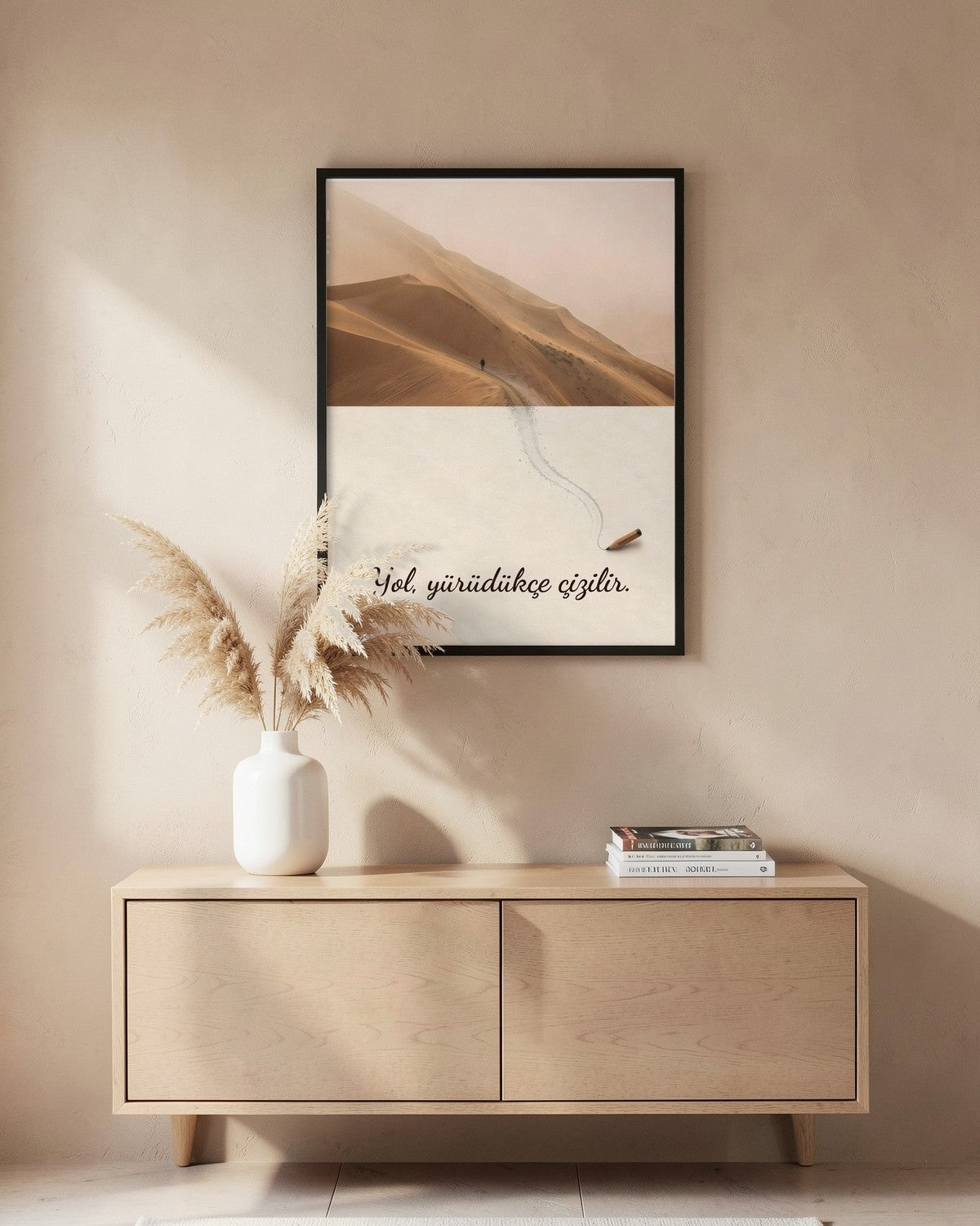 Siyah çerçeveli "Yol, yürüdükçe çizilir." posteri, bej duvarda, pampas otu ve kitaplarla süslü ahşap dolabın üstünde.